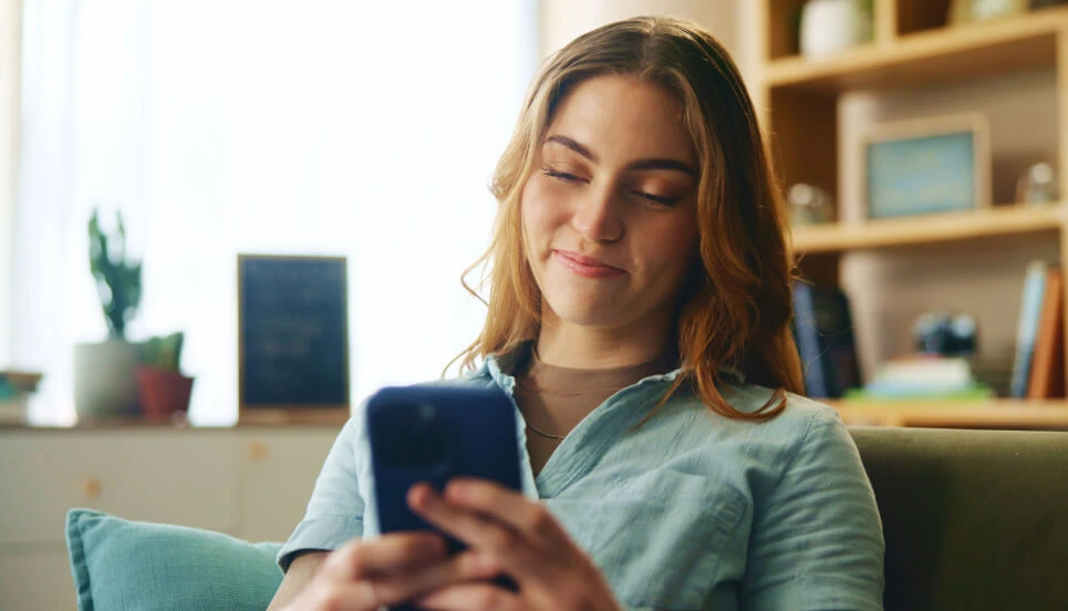 A woman sits on a couch using a smartphone, smiling slightly. Shelves with books and plants are in the background.
