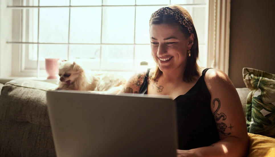 Woman smiling at a laptop on a couch, with a small dog resting beside her, near a window.