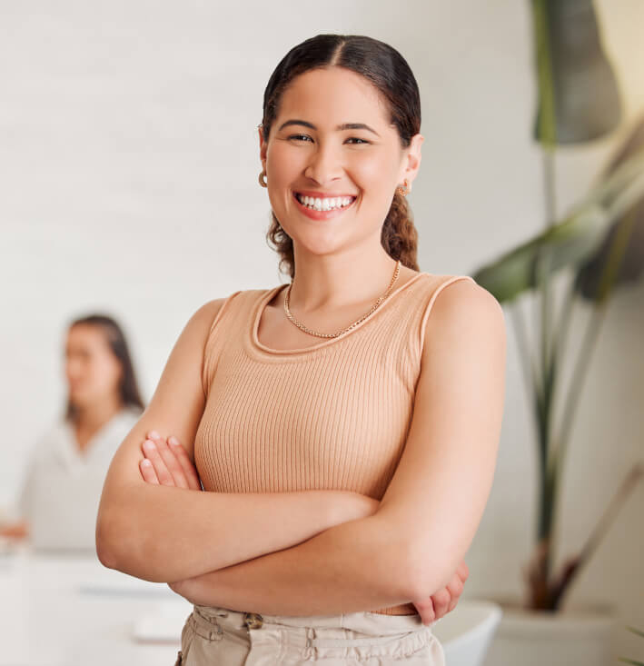 A woman stands smiling with arms crossed, wearing a beige top. Another person sits blurred in the background. Large green plant leaves are visible on the right.