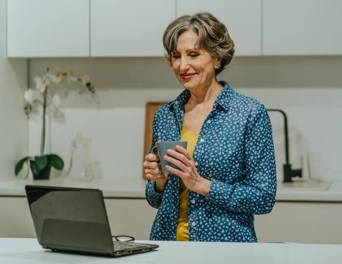 A woman in a blue shirt holds a mug while looking at a laptop in a kitchen.