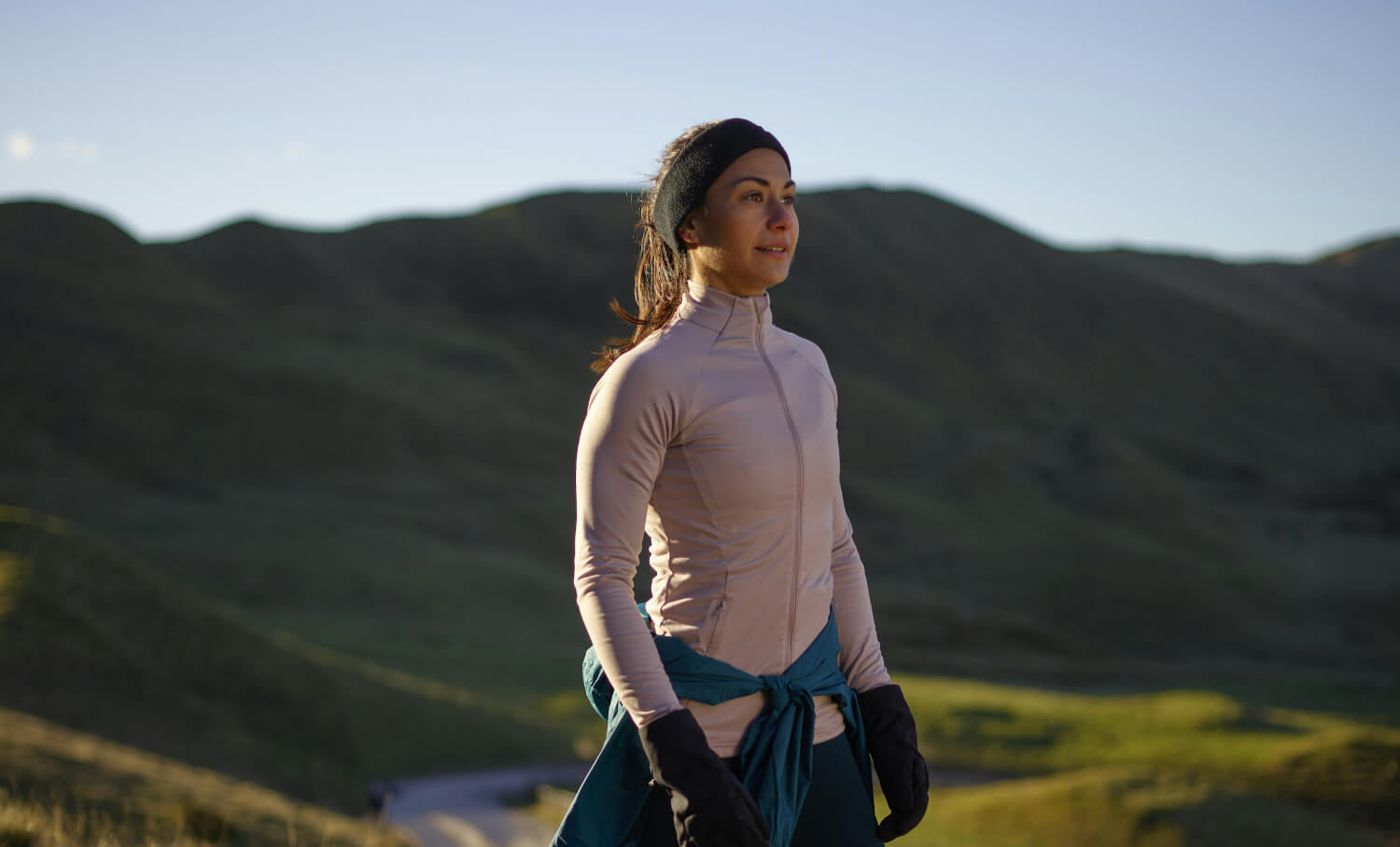 A person wearing a jacket and gloves stands on a grassy hillside with mountains in the background, looking into the distance.
