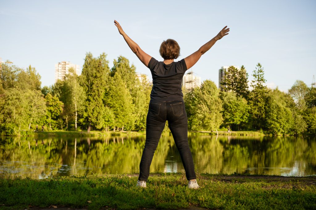 A person stands by a lake with arms raised, embracing lifestyle changes, surrounded by trees and distant buildings under a clear sky.