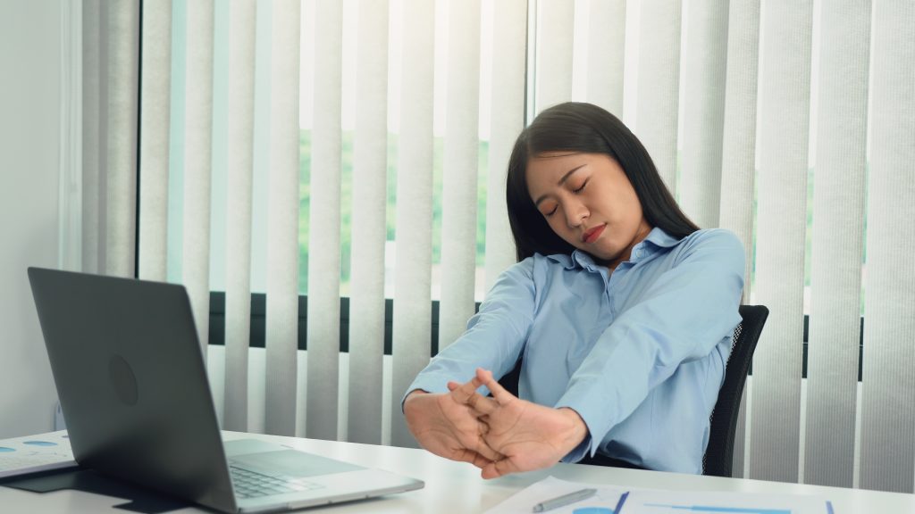 A woman in a blue shirt stretches at her desk, surrounded by a laptop and papers, as vertical blinds softly filter the light. Embracing moments of self-care amidst the digital eye strain of modern healthcare enhances her mental wellbeing.