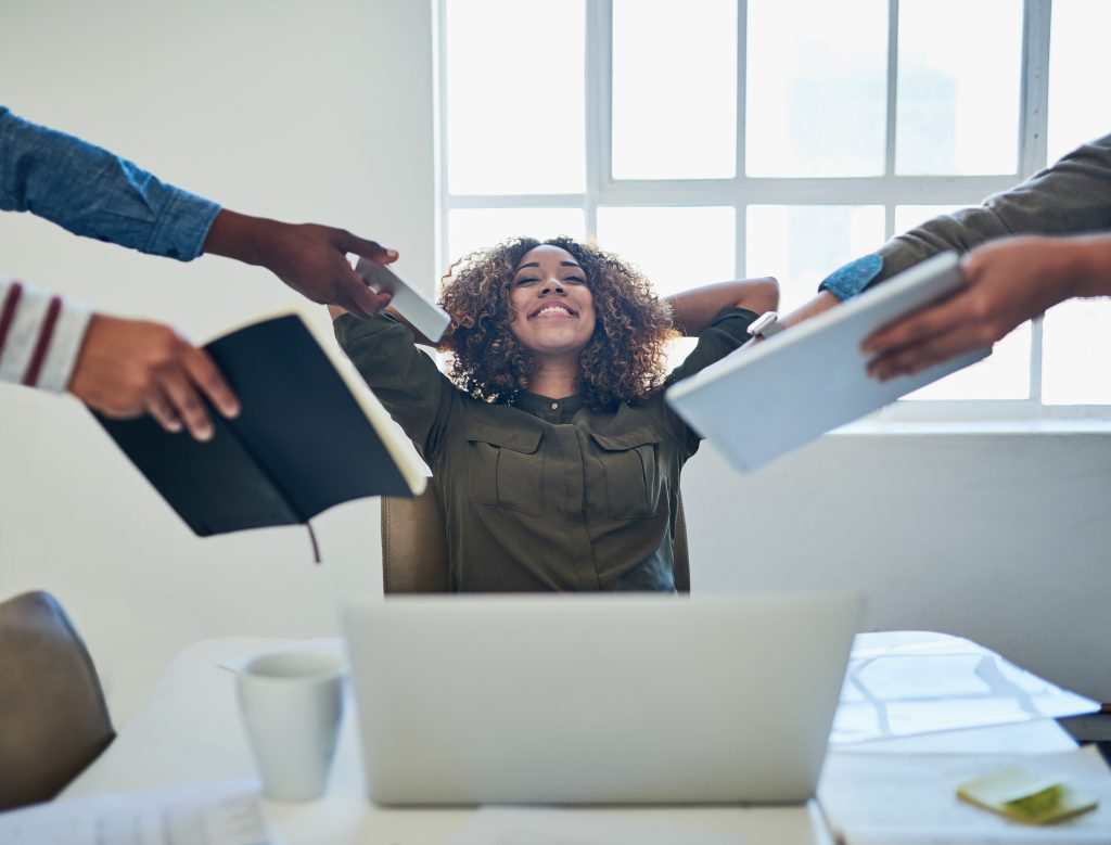 A woman at a desk stretches with her arms behind her head, surrounded by others holding notebooks and documents, finding professional support amidst work-related stress.