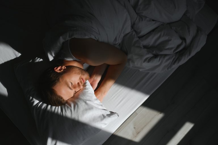 A man sleeps peacefully in bed, wrapped in white sheets, with sunlight streaming in through a window—a testament to the lifestyle changes that guarantee restful nights and make a difference in one's well-being.