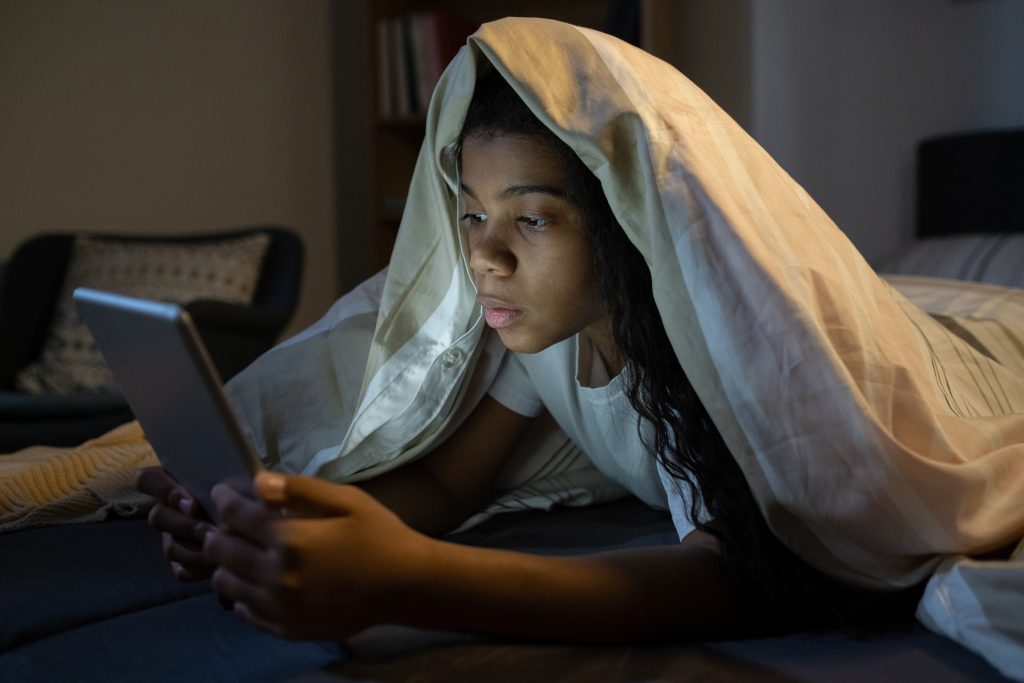 Person lying in bed under a blanket, looking at a tablet in a dimly lit room, mindful of digital eye strain and prioritizing mental wellbeing.