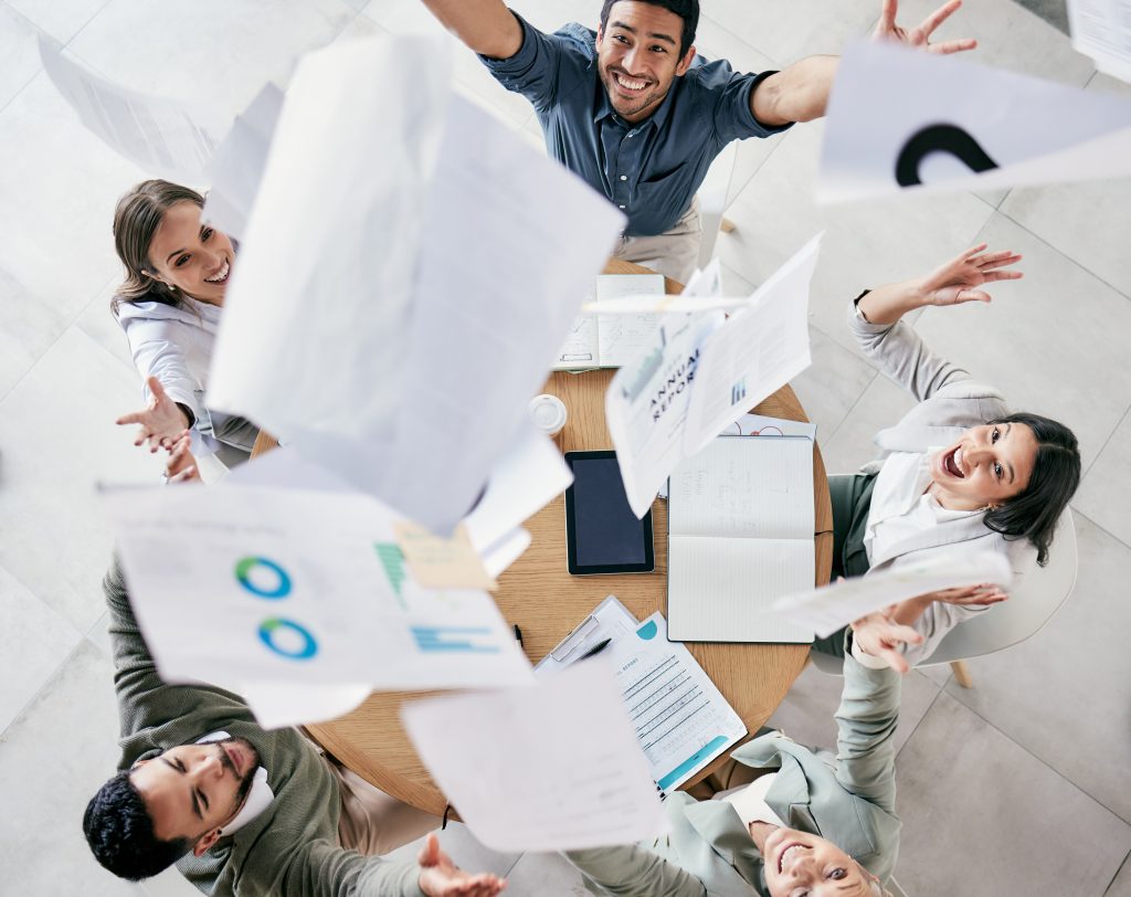 A group of four people around a table joyfully throwing papers into the air, celebrating their understanding of options. Laptops, notebooks, and documents are scattered across the table in this lively moment free from work-related stress.