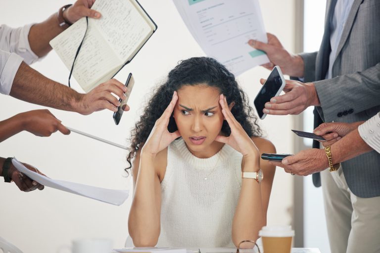 A woman sits at her desk, looking stressed as people surround her with papers, phones, and notebooks. In this moment of work-related stress, professional support offers a path to understanding your options and finding balance amidst the chaos.