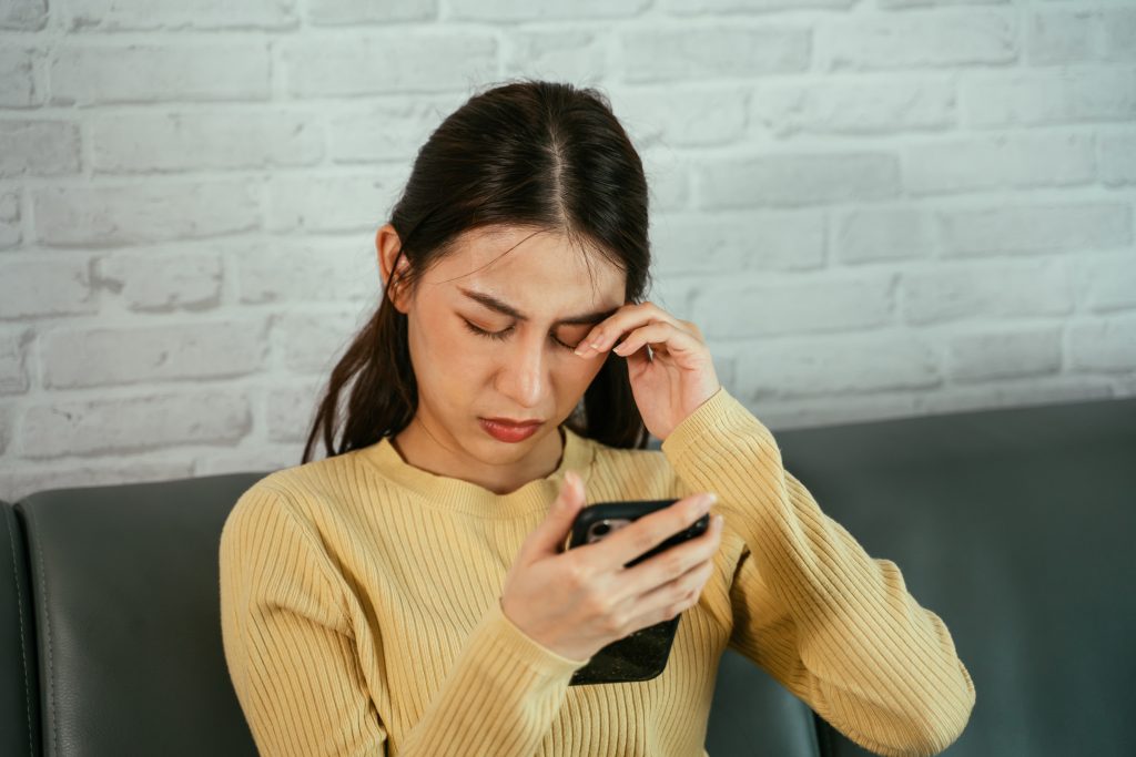 A woman in a yellow sweater looks distressed, likely experiencing digital eye strain, as she sits on the couch. Holding a smartphone in one hand and rubbing her eye with the other, her mental wellbeing seems affected by constant screen exposure.
