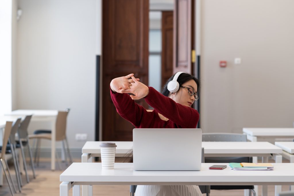 A person wearing headphones stretches in front of a laptop in a classroom, perhaps contemplating work-related stress. A coffee cup steams beside open notebooks on the table, providing an atmosphere of focus and professional support.